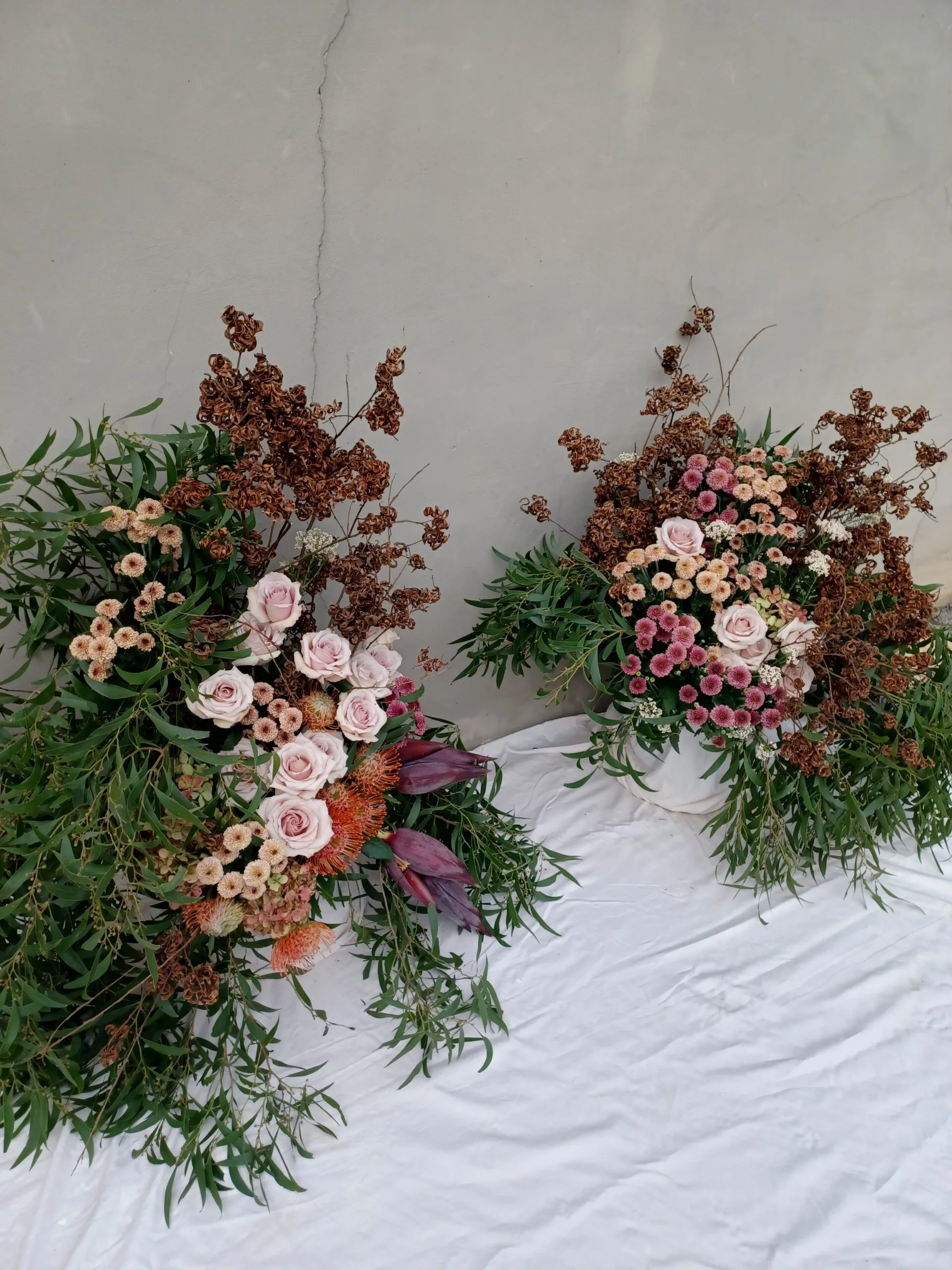 Kid Florist modern garden style wedding aisle arrangements in Matakana. The asymmetrical pair of arrangements uses roses and unusual seed pods in terracotta, rust, and blush tones. Graceful cascading greenery frames the flowers.
