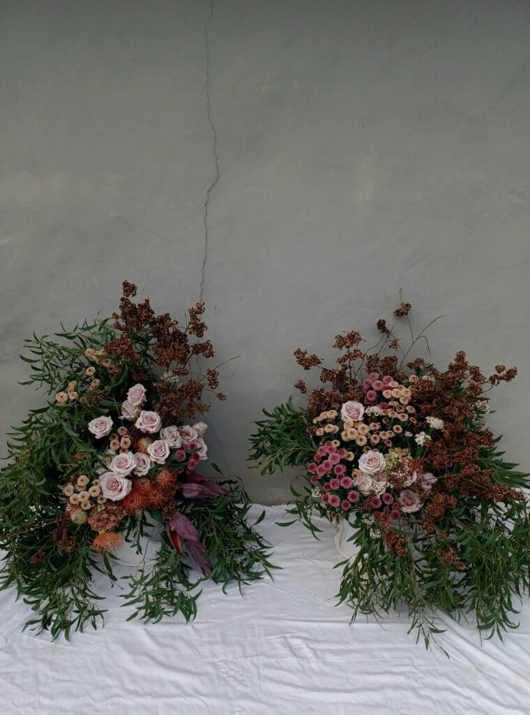 Kin Florist Matakana wedding aisle floral arrangements. Two overflowing asymmetrical arrangements feature roses and other flowers in terracotta, rust, and blush tones, and cascading greenery. The foam-free arrangements are a modern garden style, in an understated palette.
