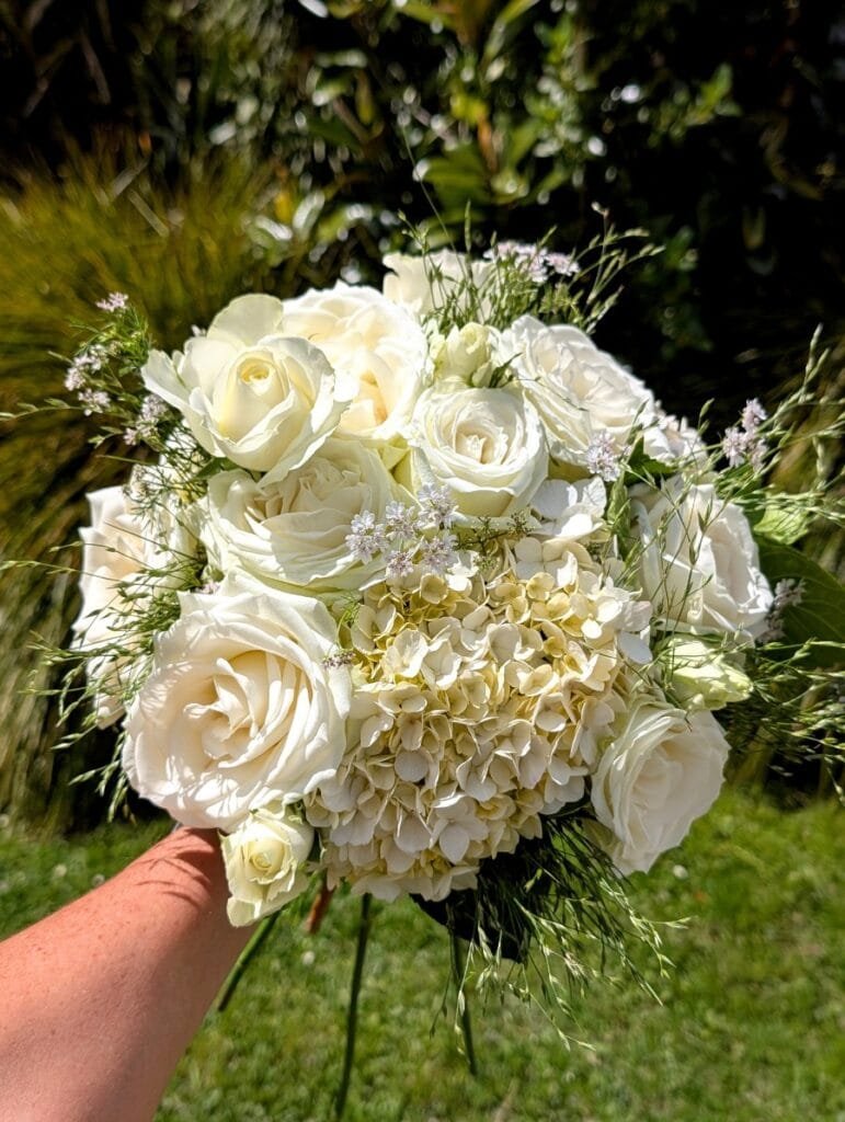 A classic white wedding bouquet by Kin Florist. It features white roses, white hydrangea, textural grass, and delicate accent flowers. The bouquet is made from all local and homegrown flowers.