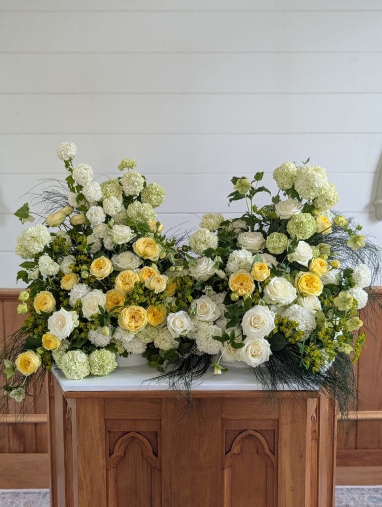 An abundant, garden style floral arrangement by Kin Florist. It features 100% local flowers - white roses, yellow garden roses, and fluffy snowballs. At St. Andrews Church it compliments the warm wood beautifully. It was later repurposed to The Farmers Daughter, where it enhanced the casual coastal vibe.